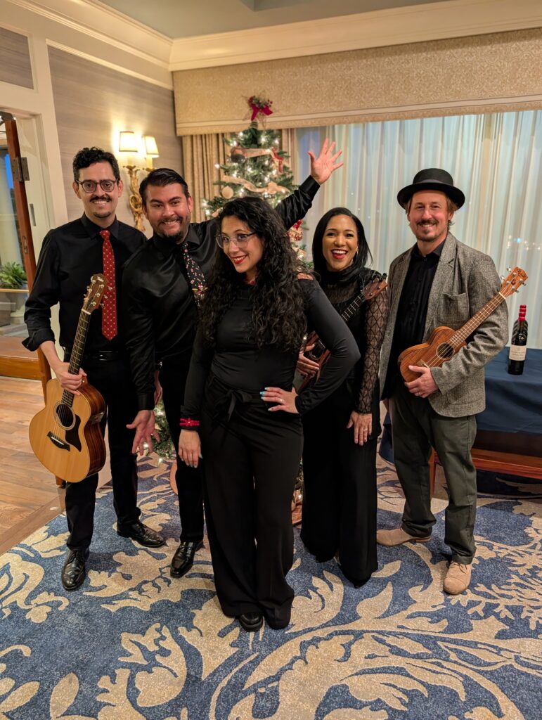 Five performers posing with acoustic instruments beside a Christmas tree